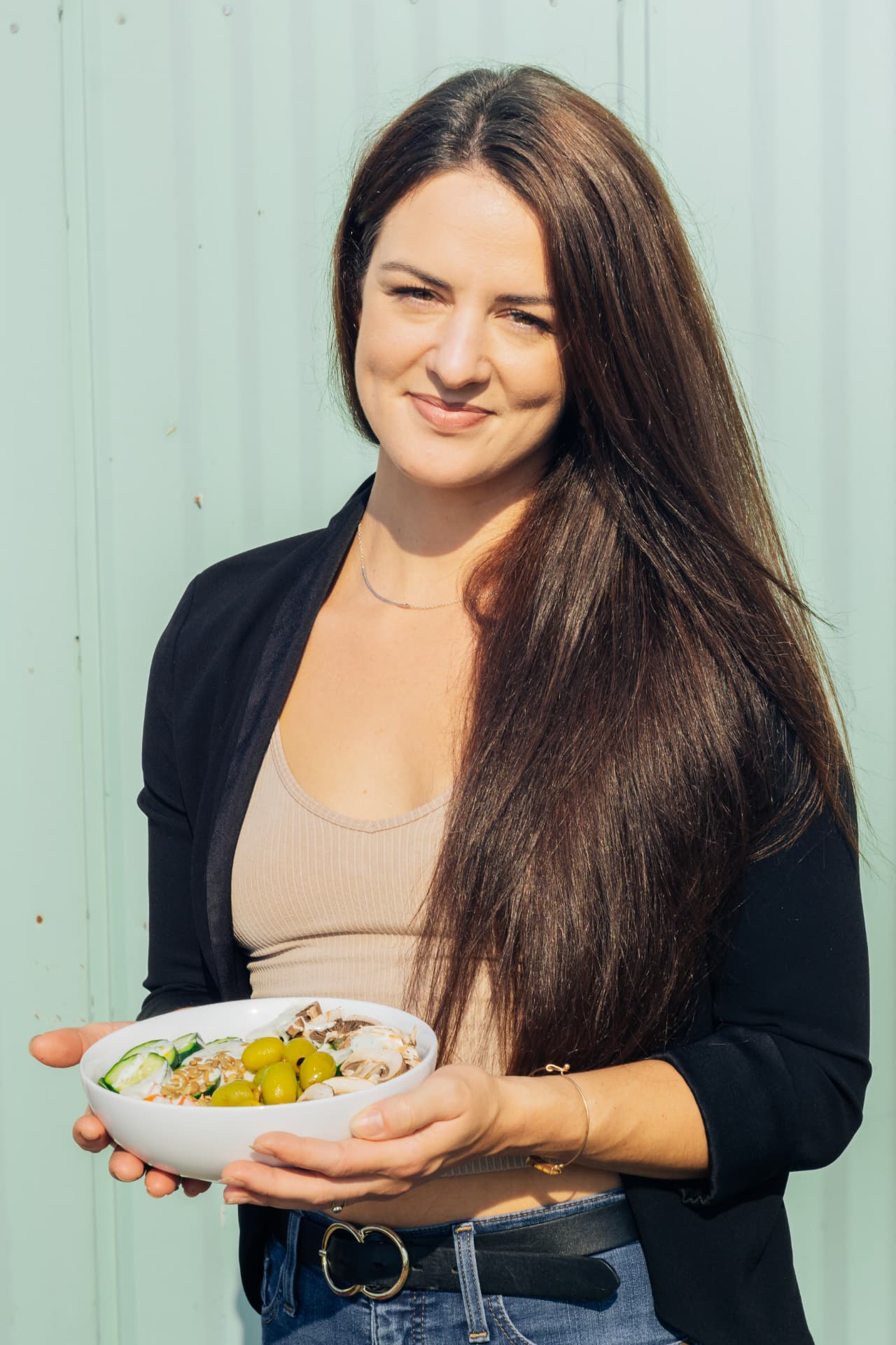 Ceylon Rose in a black blazer smiling and holding a bowl of fresh vegetables in front of a sage-green wall