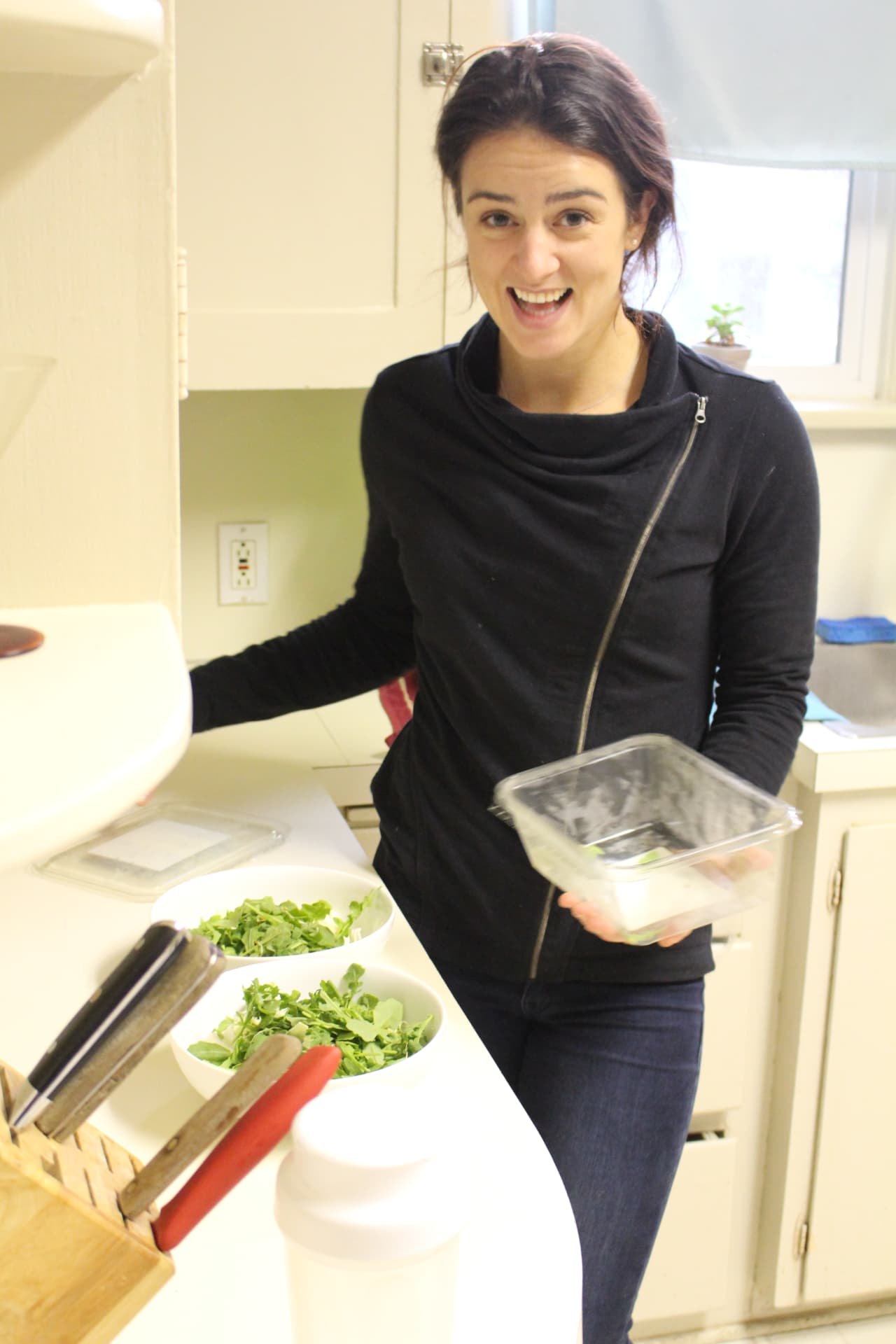 Ceylon Rose smiling behind her kitchen counter holding a food container, bowls of greens beside her
