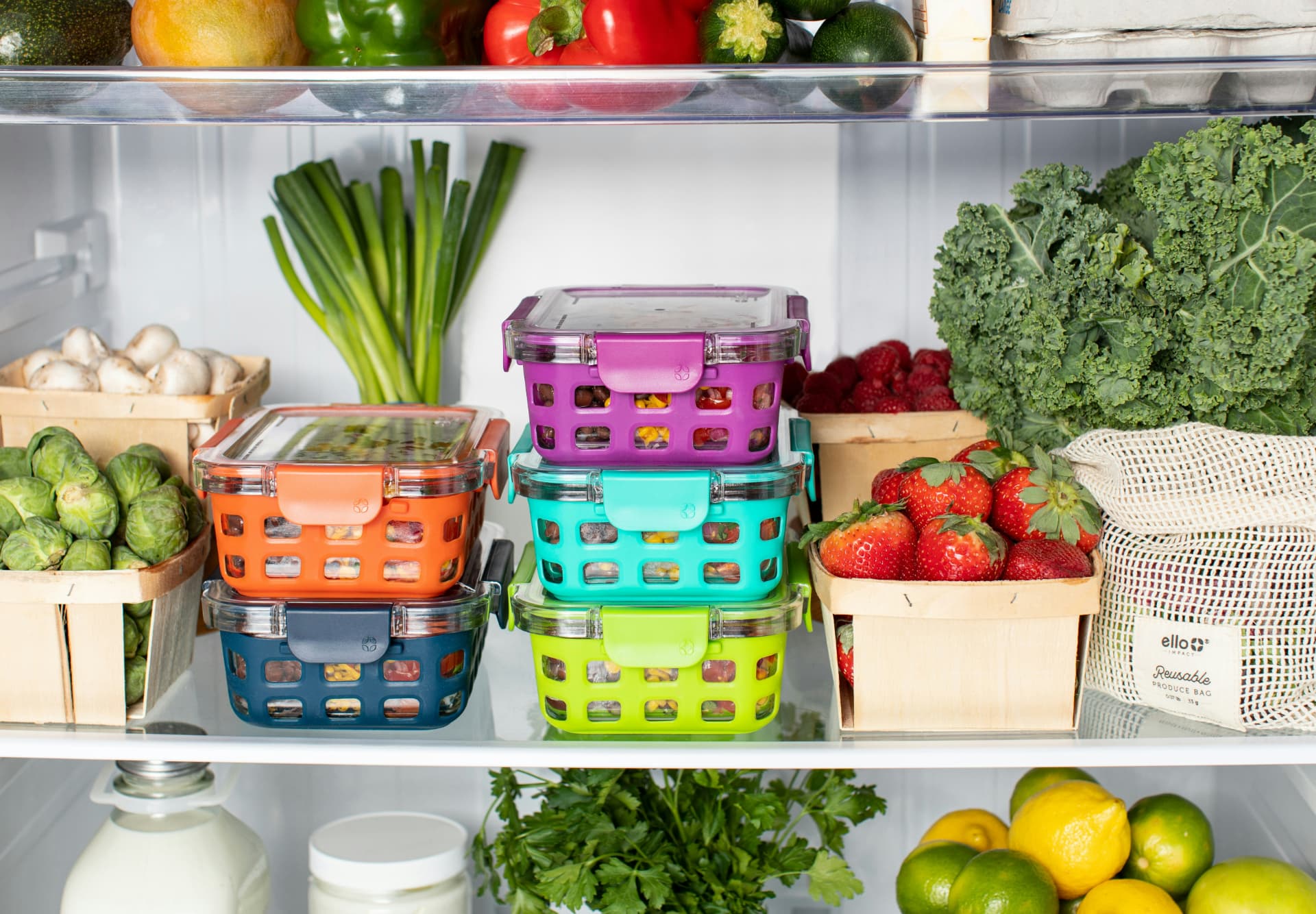 An open refrigerator with colorful glass prep containers and baskets of mushrooms, Brussels sprouts, raspberries and strawberries, leafy greens, lemons and limes
